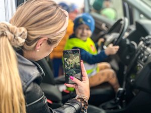 Na zdjęciu widać kobietę, odwróconą tyłem do fotografa, która trzyma w ręku telefon i robi zdjęcie dziecku siedzącemu w samochodzie. Na pierwszym planie znajduje się głowa kobiety oraz telefon. W tle widać dziecko.