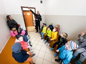 Zdjęcie przedstawia funkcjonariusza policji oraz grupę dzieci znajdujących się w pomieszczeniu dla osób zatrzymanych. Policjant prezentuje pomieszczenie.