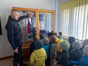 Zdjęcie przedstawia umundurowanego funkcjonariusza prezentującego sztandar Komendy Powiatowej Policji w Leżajsku, który znajduje się za szybą. Na zdjęciu widać grupę dzieci.