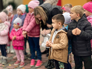 Na zdjęciu widoczne są dzieci, które powtarzają za policjantem prawidłowe przyjęcie postawy na tak zwanego żółwia. Na pierwszym planie znajduje się chłopczyk, za nim widać zamazane sylwetki dzieci.