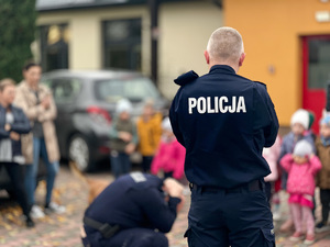 Na pierwszym planie widać dwóch umundurowanych policjantów. Jeden z nich stoi, drugi z nich jest w trakcie przyjmowania pozy na tak zwanego żółwia. W tle widać samazane sylwetki dzieci i osób dorosłych. Funkcjonariusze są odwróceni tyłem do fotografującego.