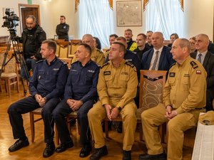 Na zdjęciu widać grupę osób znajdującą się w pomieszczeniu budynku Muzeum Ziemi Leżajskiej. W pierwszym rzędzie siedzi 4 osoby - dwóch funkcjonariuszy Policji i dwóch funkcjonariuszy Państwowej Straży Pożarnej. Ubrani oni są w mundury służbowe. Z tyłu za nimi siedzi także cztery osoby - dwóch funkcjonariuszy Policji i Państwowej Straży Pożarnej oraz dwóch mężczyzn ubranych w garnitury. Za nimi, na dalszym planie widać inne osoby biorące udział w wydarzeniu. Po lewej stronie kadru widać operatora wraz z kamerą, która stoi na statywie.