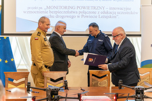 Na zdjęciu widać czterech mężczyzn, w tym dwóch umundurowanych funkcjonariuszy Policji oraz Państwowej Straży Pożarnej. Funkcjonariusz Policji wraz z mężczyzną w garniturze znajdują się na drugim planie i podają sobie ręce. Dwóch pozostałych mężczyzn znajduje się na pierwszym planie. Mężczyzna stojący po prawej stronie trzyma w ręku niebieską teczkę. Na stole przed nimi stoją dwa drony, które na zdjęciu są zamazane.