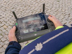 Na zdjęciu widać zbliżenie na aparaturę sterującą dronem. Aparatura trzymana jest Policjanta, który ubrany jest w mundur. Na zdjęciu najostrzejszym elementem jest aparatura.