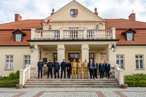 Na zdjęciu znajduje się trzynaście osób ustawionych w rzędzie. Stoją bezpośrednio przy wejściu do Muzeum Ziemi Leżajskiej. Wśród tych osób znajdują się umundurowani funkcjonariusze Policji i Państwowej Straży Pożarnej. osoby zwrócone są frontem do aparatu.