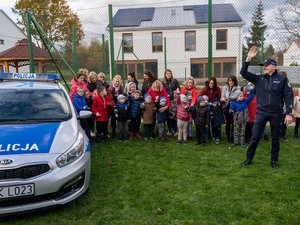 Na zdjęciu, po lewej stronie widoczny jest oznakowany radiowóz policji. Stoi zaparkowany na trawniku. Obok niego stoi grupa dzieci wraz z opiekunami. Po prawej stronie widoczny jest umundurowany funkcjonariusz Policji, który ma uniesioną swoją prawą rękę. Wszyscy stoją frontem do obiektywu kamery.