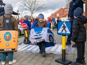 Na zdjęciu, na pierwszym planie widoczny jest chłopiec, który ma założony na sobie wykonany z materiału radiowóz. Chłopiec patrzy w kierunku aparatu. Po jego obu stronach widoczne są imitacje znaków drogowych. Chłopiec przechodzi przez makietę przejścia dla pieszych. W tle widać siedzi i opiekunów. Po lewej, jak i zarówno po prawej stronie widać sylwetki policjantów, którzy są umundurowani.