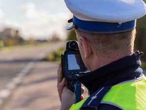 Zdjęcie przedstawia umundurowanego policjanta z urządzeniem pomiarowym prędkość. Policjant stoi tyłem do obiektywu aparatu. W tle widać drogę.