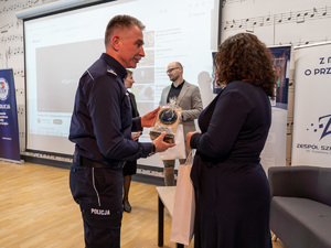 Zdjęcia przedstawiają przebieg debaty. Na zdjęciach widoczni są umundurowani policjanci oraz prelegenci. Debata społeczna odbyła się w Zespole Szkół Licealnych im. Bolesława Chrobrego. Na wybranych kadrach widoczna jest tłumnie zgromadzona na sali koncertowej młodzież.