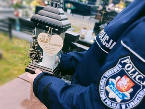 Na zdjęciu policjant w umundurowaniu służbowym trzyma w dłoni znicz w tle cmentarz.