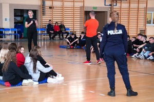 Na zdjęciu instruktor samoobrony razem z policjantką wygłaszającą prelekcję oraz policjantka w umundurowaniu służbowym. W tle uczestnicy spotkania.