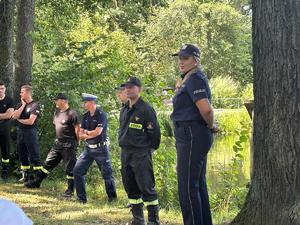 Na zdjęciu policjanci i strażacy w umundurowaniu służbowym na tle lasu i zalewu.