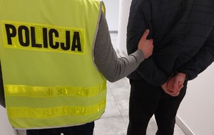 Na zdjęciu plecami policjant w kamizelce odblaskowej i osoba z kajdankami na rękach trzymanych z tyłu.