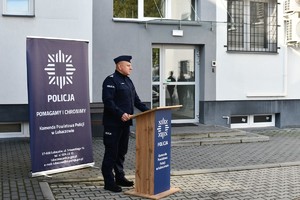 Komendant Powiatowy Policji w Lubaczowie, w umundurowaniu służbowym stoi przy mównicy z logo komendy i przemawia, w tle roll-up oraz budynek komendy.