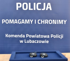 Na tle napisu &quot;Policja POMAGAMY I CHRONIMY Komenda Powiatowa Policji w Lubaczowie&quot; dwa kluczki do radiowozu leżące na tacy.