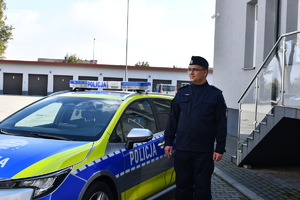 Umundurowany policjant stojący przy radiowozie w tle budynek.