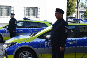 Dwóch umundurowanych policjantów przy radiowozach w tle budynek komendy.