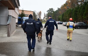 Trzech policjantów, pies służbowy i strażak idą na miejsce zdarzenia, w tle budynki i dzieci.