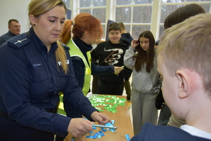 Na sali gimnastycznej uczniowie przy stole z odblaskami policjanta rozdaje odblaski.