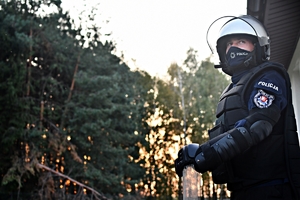 Na tle drzew policjant umundurowany w zestawie przeciwuderzeniowym.