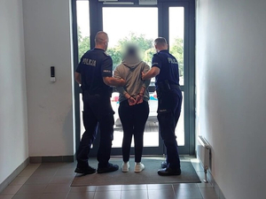 Dwóch umundurowanych policjantów prowadzi kobietę, która ma założone kajdanki na ręce trzymane z tyłu.