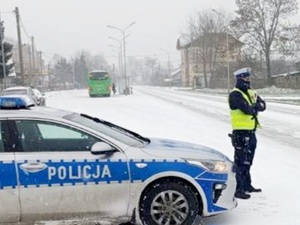 policjant stoi przy drodze za nim osobowy radiowóz w tle przystanek autobusowy i autobus zimowa aura