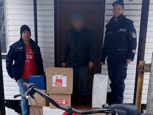 policjant i dwóch mężczyzn stoją przed domem z rowerem obok ułożone kartonowe pudełka z napisem szlachetna paczka