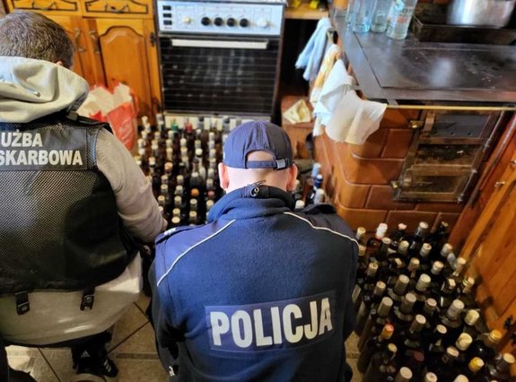 w pomieszczeniu policjant i funkcjonariusz służby celno skarbowej widok od strony pleców, przed nimi ułożone butelki wypełnione brązową cieczą