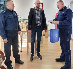 mężczyzna stoi pomiędzy policjantami którzy wręczają mu dyplom w tle godło Polski