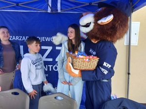 policyjna maskotka lew podczas pikniku rodzinnego trzyma w rękach kosz ze słodyczami obok niego młodzież