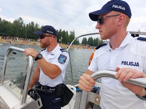 policjanci na łodzi motorowej na wodzie patroluja teren zbiornika wodnego na brzegu którego wypoczywają osoby