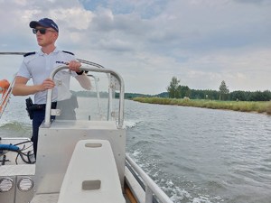 policjanci na łodzi motorowej na wodzie patroluja teren zbiornika wodnego na brzegu którego wypoczywają osoby