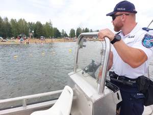 policjanci na łodzi motorowej na wodzie patroluja teren zbiornika wodnego na brzegu którego wypoczywają osoby