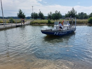 policjanci na łodzi motorowej na wodzie patroluja teren zbiornika wodnego na brzegu którego wypoczywają osoby