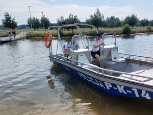 policjanci na łodzi motorowej na wodzie patroluja teren zbiornika wodnego na brzegu którego wypoczywają osoby