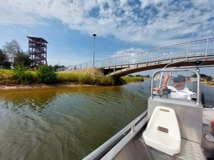 policjanci na łodzi motorowej na wodzie patroluja teren zbiornika wodnego na brzegu którego wypoczywają osoby