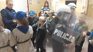 Spotkanie z dziećmi z Gromady Zuchów ze Szkoły Podstawowej w Grochowcach. Dzieci na terenie  Komendy Miejskiej w Przemyślu ustawione w grupie słuchają prelekcji policjantki i ubierają elementy umundurowania i sprzętu taktycznego policjanta