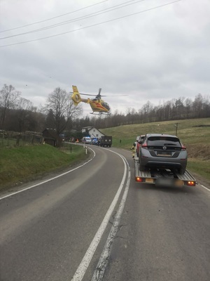 zdjęcia ze zdarzenia drogowego w Boguszówce. na zdjęci na drodze stoi samochód z lawetą , widać lecący na d drogą  helikopter LPR, w tle miejsce zdarzenia