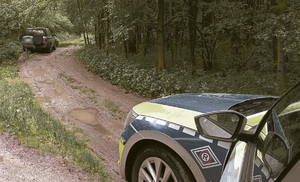 Na zdjęciu widać oznakowany radiowóz policyjny na błotnistej drodze z kałużami. W oddali stojący pojazd marki land rower oraz dużo drzew liściastych.