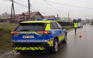 Zdjęcie z miejsca zdarzenia. Na fotografii widać oznakowany radiowóz stojący na drodze. W tle widoczne uszkodzone auto również stojące na drodze.